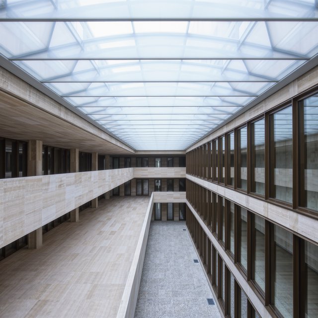 A soaring civic atrium in contemporary Italian Rationalist style pairs pale travertine, bronze mullions, and terrazzo beneath a glazed roof; cool morning light falls from above, viewed from a mezzanine, with aligned bays and credible supports. — Kling v3 Omni