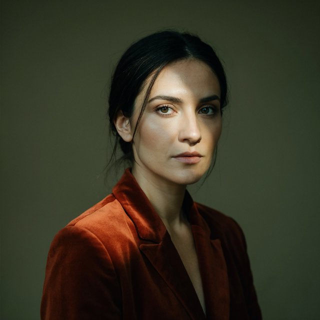Her guarded gaze catches a solitary beam in a low-key fashion portrait: an adult woman aged 27–35 wears a rust-colored velvet blazer, the rest of her face falling into shadow against a deep olive backdrop. — Ideogram v4