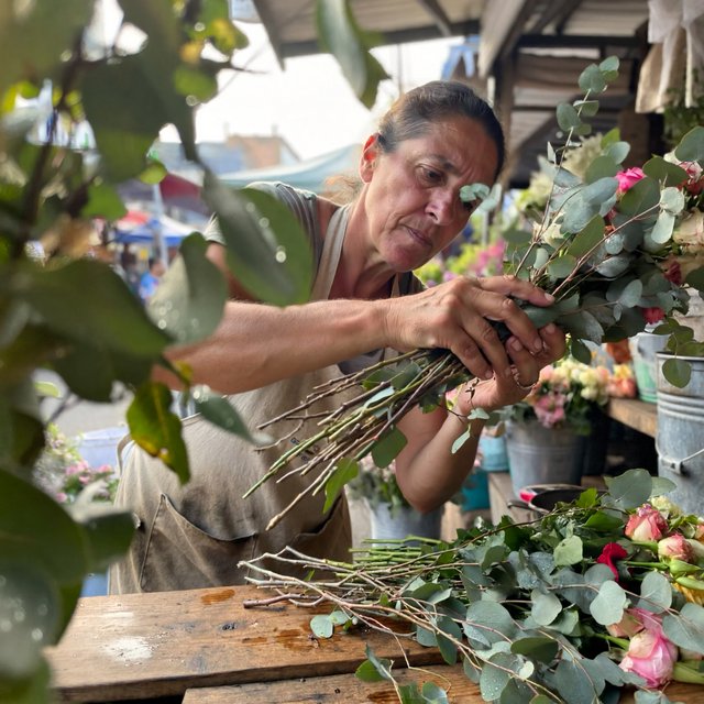 Candid documentary photograph of an adult florist arranging loose stems at an outdoor market stall, soft early-morning side light, 50mm lens with shallow depth of field, tiny water droplets beading on the cut leaves. — ImagineArt 2