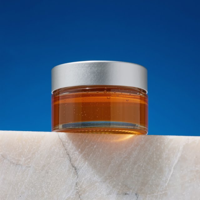 A squat unbranded honey jar with clear glass walls and a plain brushed-aluminum lid rests on honed pale limestone; broad diffused key light and warm rim light shape the commercial product image against a seamless cobalt-blue background, with no lettering. — Model 10