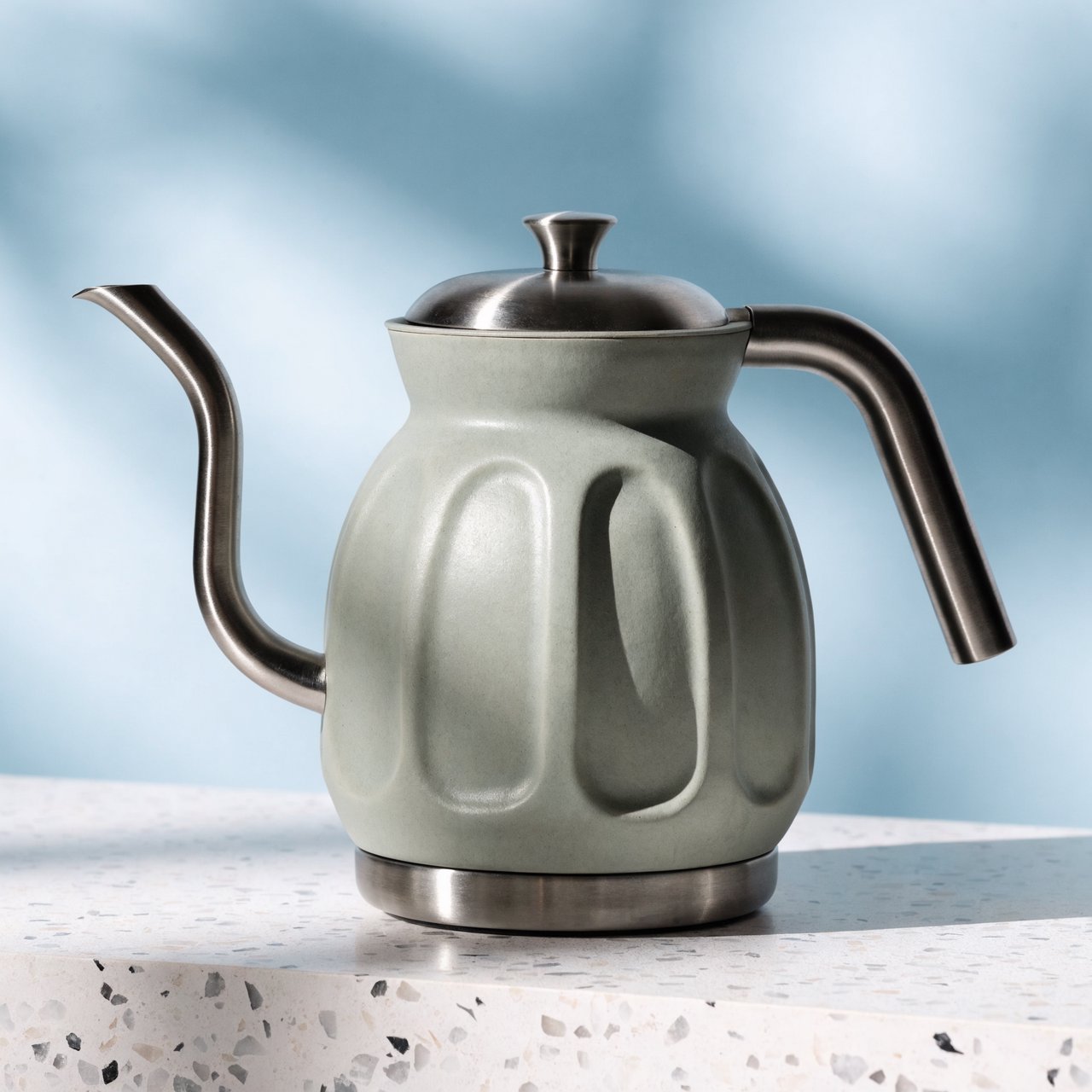 Commercial product photograph of an unbranded electric kettle made from matte sage ceramic and brushed stainless steel, resting on a white terrazzo surface against a pale blue background, with soft window light from the left and a delicate grounding shadow. — Recraft v4 Pro