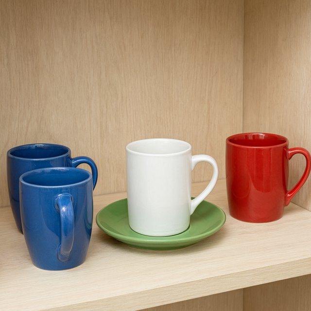 Exactly four ceramic mugs sit on a pale wooden shelf: two blue mugs on the left, one white mug in the center, and one red mug on the right. A green saucer is beneath the center mug, nothing else. — Qwen Image 3.0 Pro