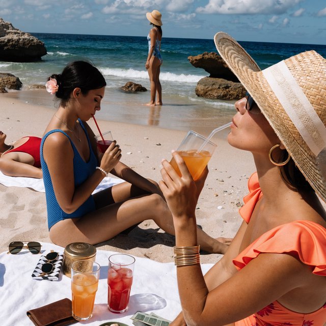 A realistic scene of women in one-piece swimsuits on the beach, with a peaceful vibe, orange and red drinks nearby, high-quality items nearby, a bit of money also nearby, and many angles. — Recraft v4 Pro