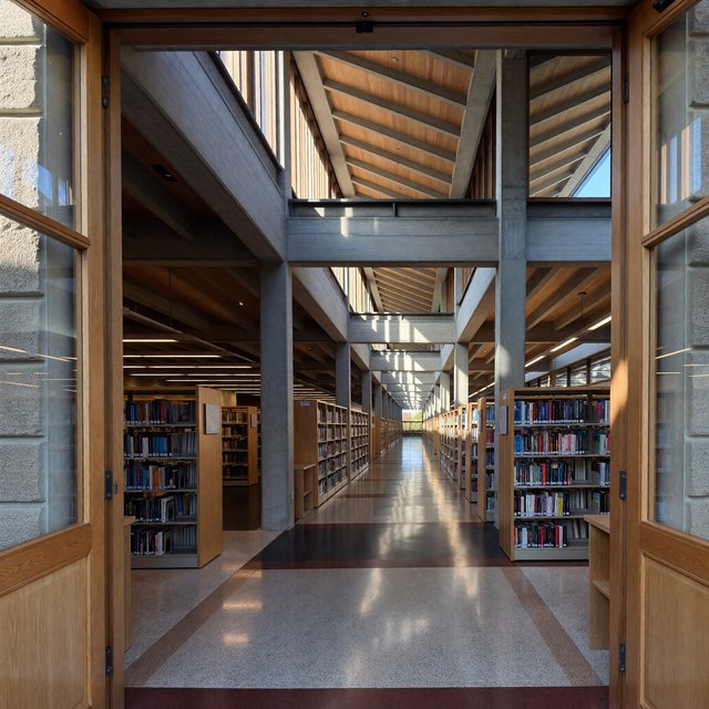 Interior of a mid-century modernist public library, with pale oak shelving, exposed concrete columns, terrazzo flooring, and a broad clerestory. Diffuse morning light crosses the reading hall, viewed from the entrance down its central aisle, with precise lines and plausible structure. — ImagineArt 1.5 Pro