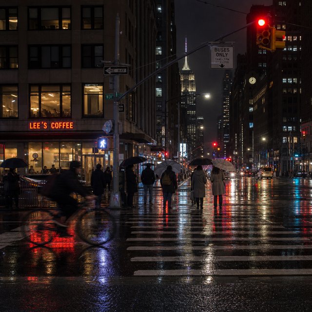 A rain-soaked city crosswalk at night seen from across the street, umbrellas, reflections of traffic lights on wet asphalt, slight motion blur on a passing cyclist. — GPT Image 2