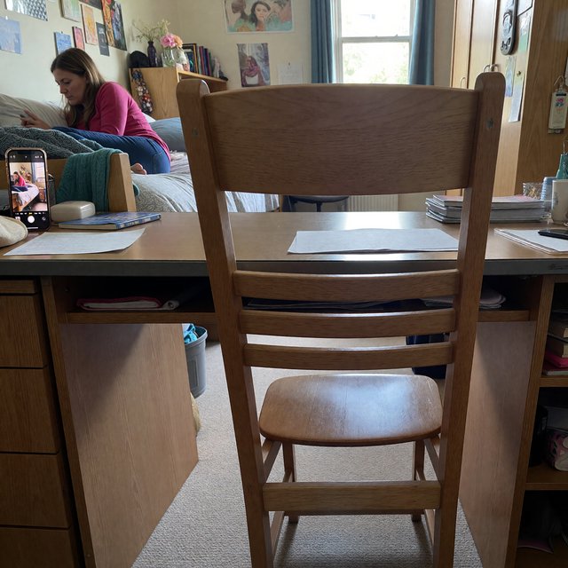 A dorm room belonging to an adult woman, photographed with a cell phone. The camera is sitting on a desk facing the empty chair and the rest of the room. — ImagineArt 1.5 Pro