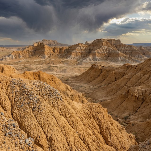 Wind-driven dust devils spiral across ochre badlands beneath a storm-clearing late-afternoon sky; pebble-studded clay ridges and tiny erosion grooves sharpen nearby, while layered mesas recede through veils of rain. — Qwen Image 3.0 Pro