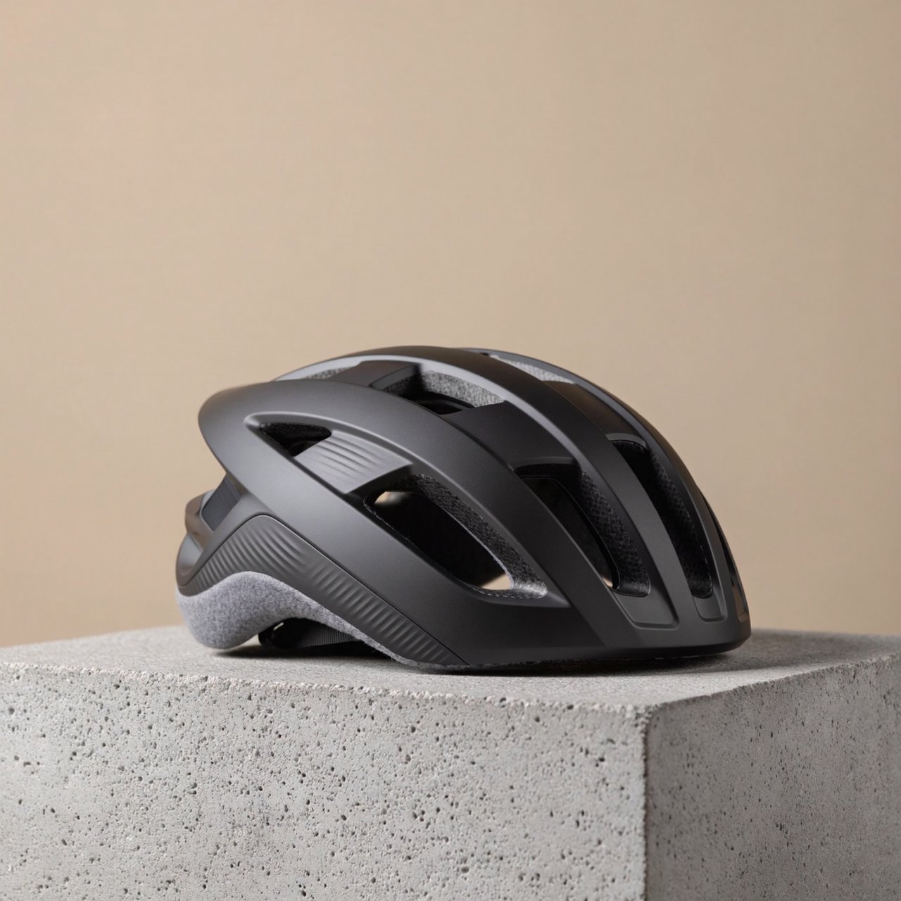 Commercial product photograph of an unbranded folding bicycle helmet made from matte charcoal polymer with soft gray padding, resting on a honed concrete plinth against a muted sand background, illuminated by a broad overhead softbox and gentle rim light. — ImagineArt 2