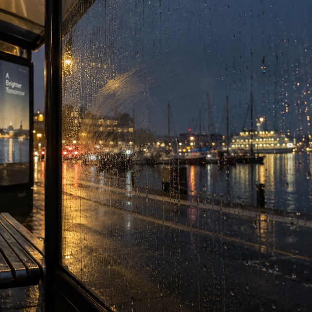 Raindrops crawl down a bus shelter pane, warping blurred harbor lights beyond. A handheld evening street snapshot in cool sodium glow, shot at 50mm with shallow depth of field, catches tiny water trails and a faint wipe mark. — GPT Image 2.5 Sunburst