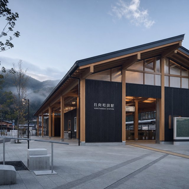 Contemporary Japanese timber train station, charred cedar cladding, pale concrete platform, broad pitched roof with exposed beams and translucent clerestories, cool morning light, viewed obliquely from the quiet approach plaza, precise straight lines and plausible structure. — GPT Image 2