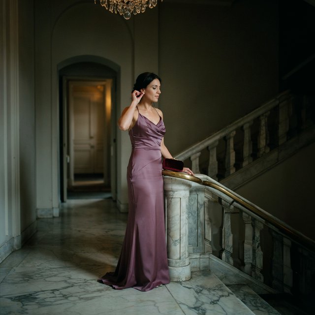 At the landing of a marble staircase, an adult woman in a plum evening gown pauses to fasten an earring, caught in warm chandelier light; polished brass rails and soft shadows lend the scene the poise of a fashion magazine. — Ideogram v4