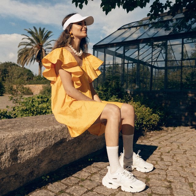 Editorial glamour photograph of an adult woman in a saffron tennis dress and white visor, resting naturally beside a glasshouse garden, flattering morning light, leafy reflections, relaxed smile, refined fashion-magazine composition. — Recraft v4