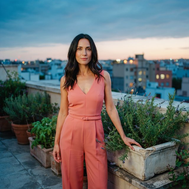 Editorial glamour photograph of an adult woman in a tailored coral evening jumpsuit on a rooftop herb garden at blue hour, resting one hand on a stone planter, flattering sunset glow, relaxed expression, city lights softly blurred behind her. — Ideogram v4.5
