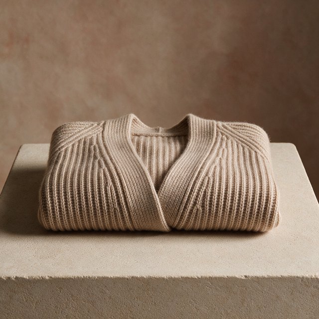 A folded unbranded merino cardigan rests on a honed limestone plinth, its chunky ribbed wool surface softly catching broad overhead diffusion and warm side bounce against a muted clay backdrop, with no tags, lettering, or graphics. — Seedream 5.0 Pro