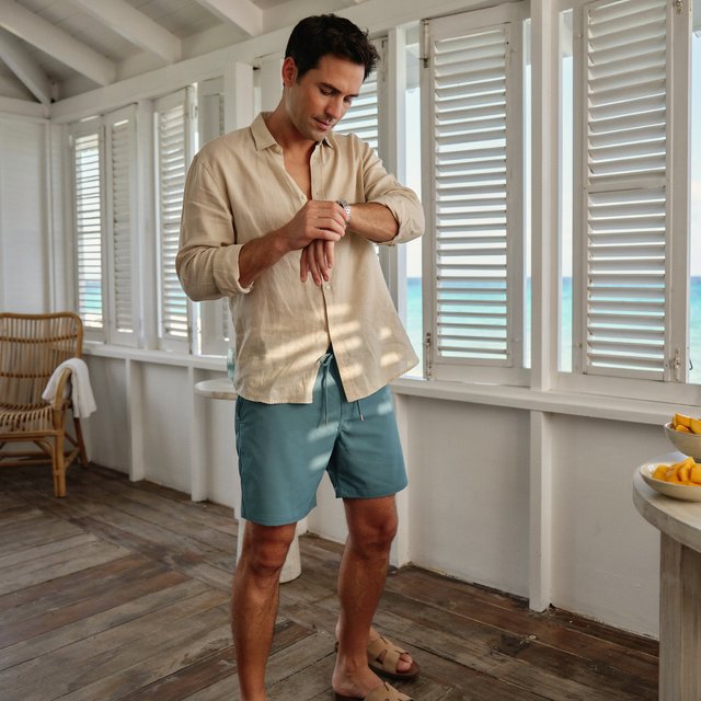 At a sunlit island veranda, an adult man in teal swim shorts and a sand-colored linen shirt is caught fastening his watch, crisp noon light and relaxed tailoring lending fashion-magazine polish. — Ideogram v4