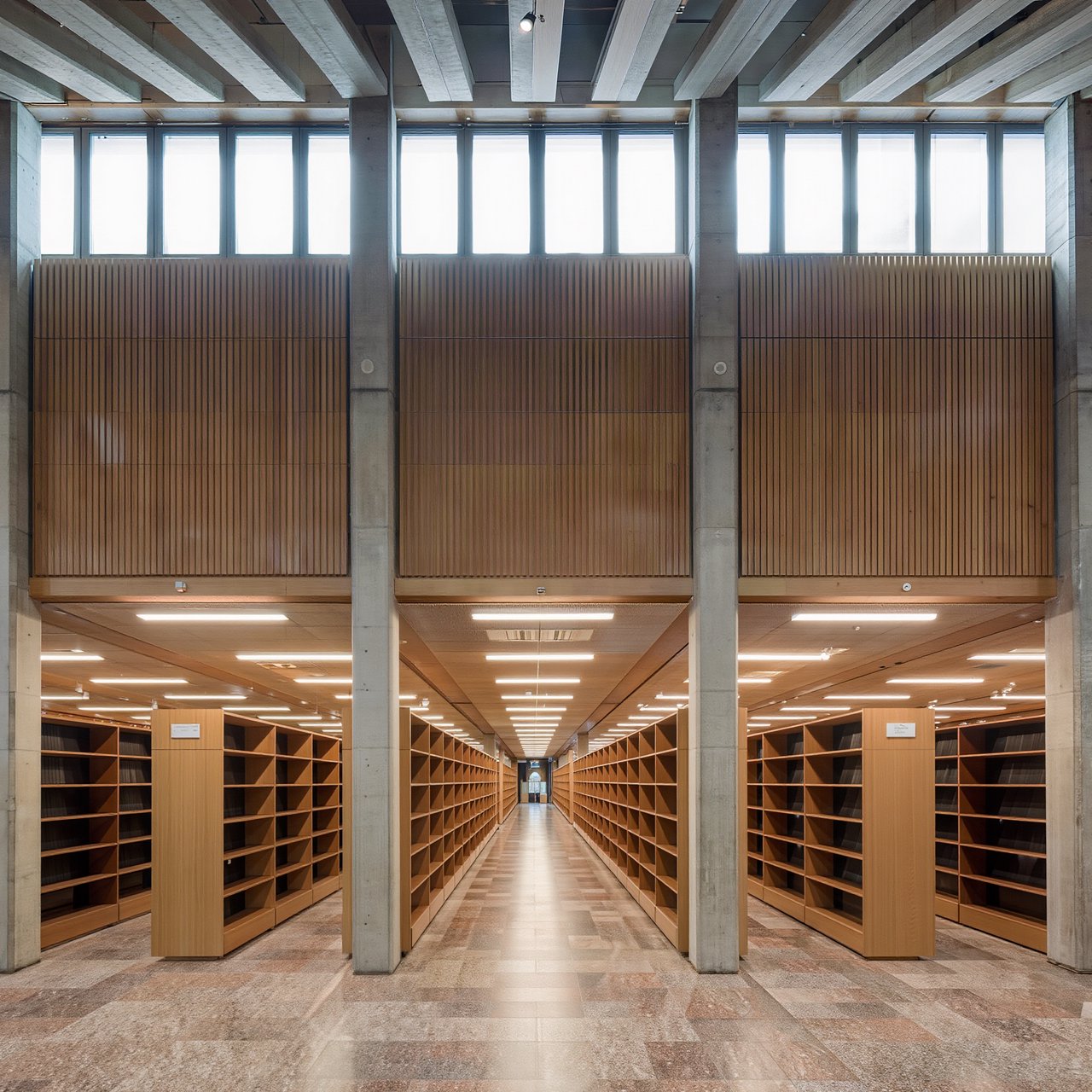 Interior of a mid-century modernist public library, with pale oak shelving, exposed concrete columns, terrazzo flooring, and a broad clerestory. Diffuse morning light crosses the reading hall, viewed from the entrance down its central aisle, with precise lines and plausible structure. — Kling v3