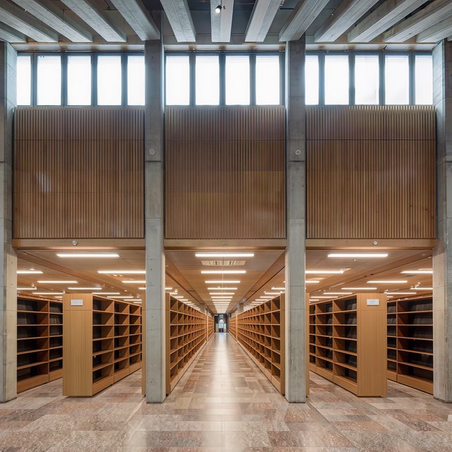 Interior of a mid-century modernist public library, with pale oak shelving, exposed concrete columns, terrazzo flooring, and a broad clerestory. Diffuse morning light crosses the reading hall, viewed from the entrance down its central aisle, with precise lines and plausible structure. — Kling v3