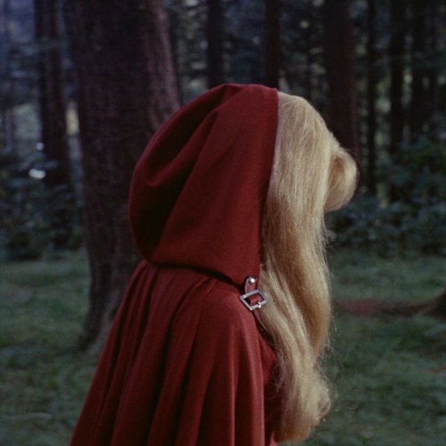Medium shot at eye level: 1960s vintage film aesthetic. An adult woman with long, blonde hair walks alone through a dark forest. She wears a red hooded cloak, with the hood pulled up and fastened at the neck by a silver buckle. Photorealistic 1960s Technicolor film grain; soft, shallow depth of field. — Krea 2 Large