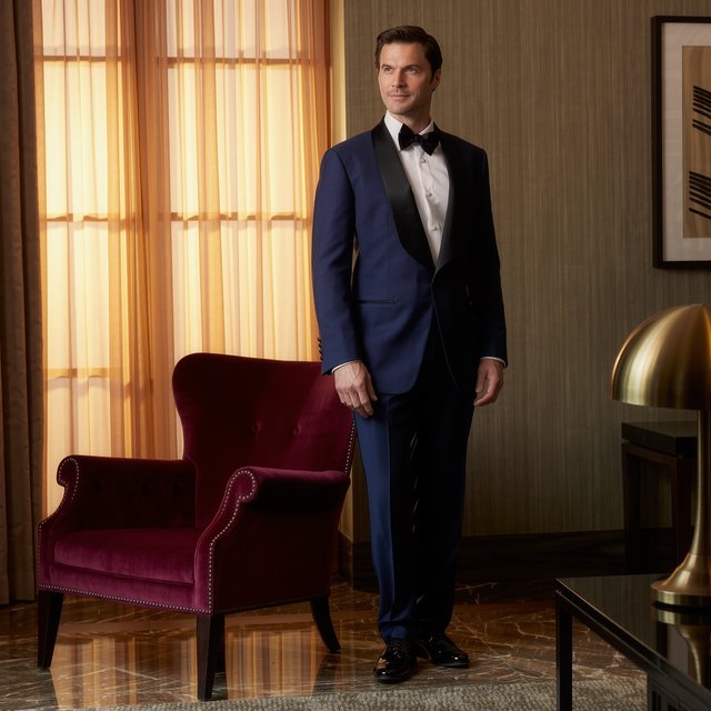 Editorial glamour photograph of an adult man in a midnight-blue tuxedo, standing naturally beside a velvet chair in a quiet hotel lounge, flattering amber window light, relaxed expression, polished magazine composition. — Kling v3 Omni