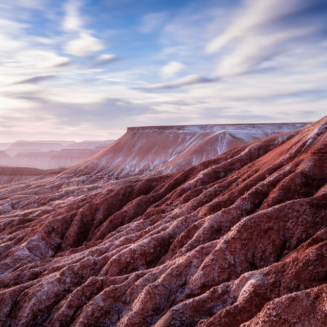 A red-sandstone badlands plateau beneath clearing winter clouds at sunrise, overnight frost silvering the gullies; cracked clay and wind-smoothed rock lead the eye across layered mesas fading into pale distance. — Krea 2 Large