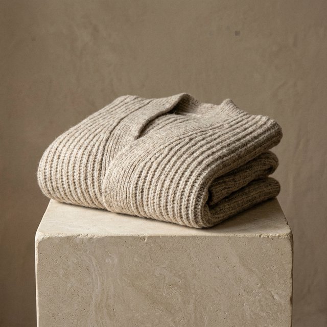A folded unbranded merino cardigan rests on a honed limestone plinth, its chunky ribbed wool surface softly catching broad overhead diffusion and warm side bounce against a muted clay backdrop, with no tags, lettering, or graphics. — Nano Banana 2.1