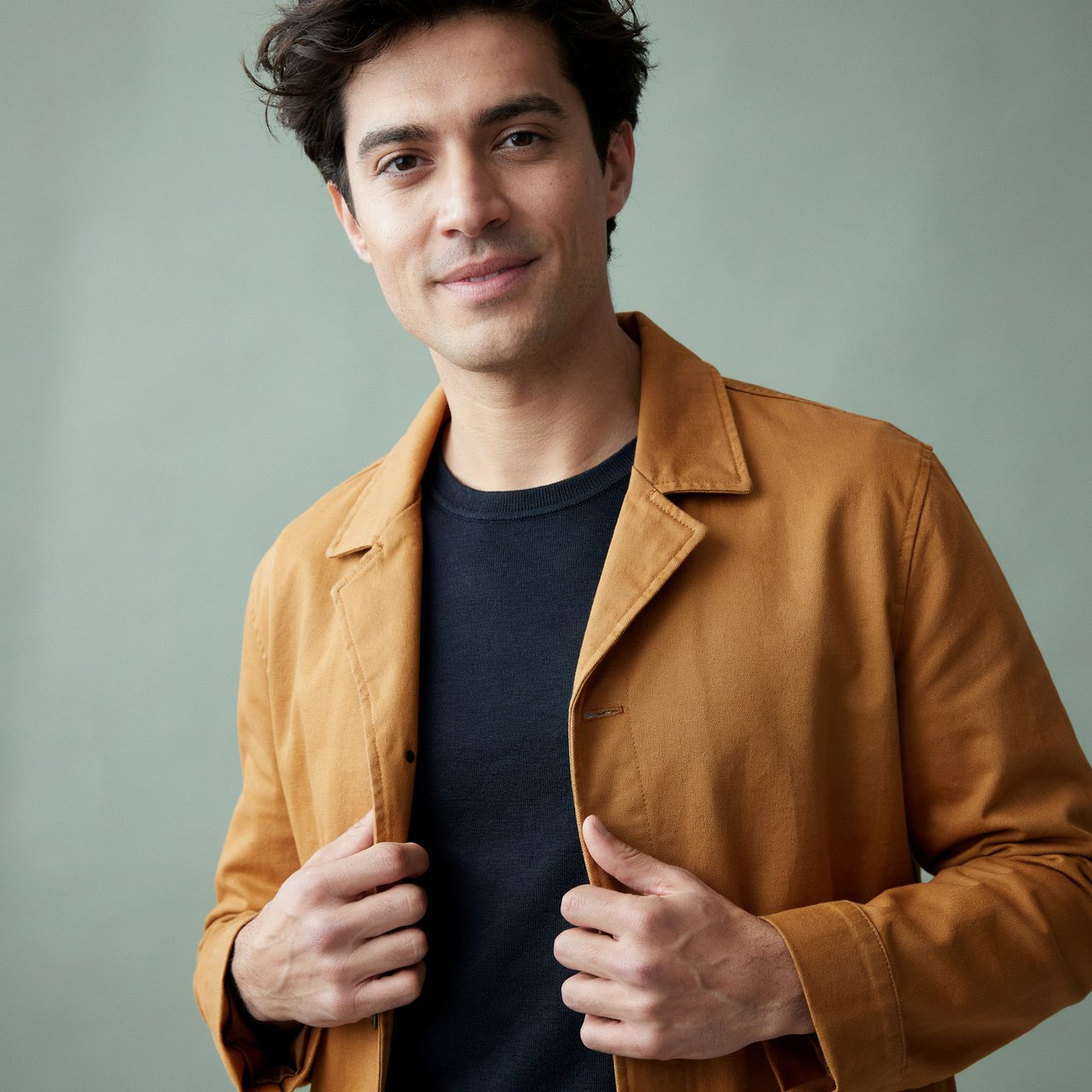 Fashion portrait of an adult nonbinary person in their late twenties wearing a structured saffron-yellow canvas jacket over a fine-gauge navy merino top, hands resting on the jacket hem. Their bright, self-assured expression contrasts with a softly lit sage-green backdrop. — Ideogram v4.5