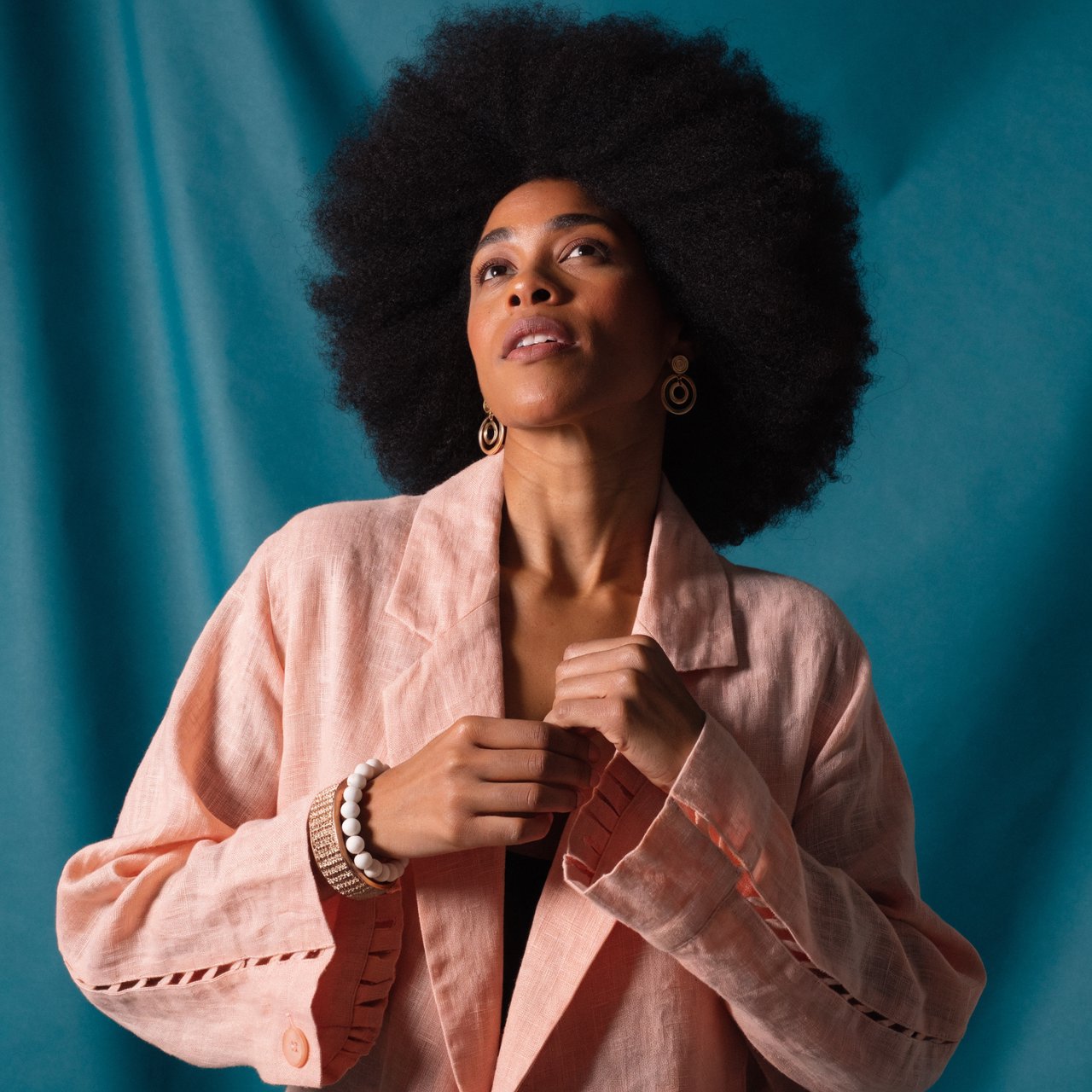 Portrait photograph of an adult woman in her early 30s wearing a pale apricot linen blazer, her hands gently smoothing its cuff. She looks quietly delighted against a deep teal paper backdrop, illuminated by soft, angled studio light. — Recraft v4 Pro