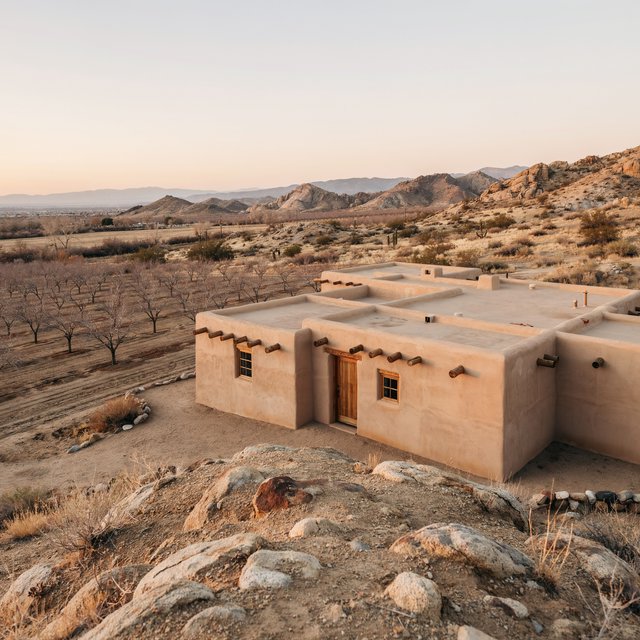 Beyond dry orchard rows, a Pueblo Revival adobe house settles into the desert foothills, with timber lintels and a muted clay roof; warm late-afternoon light, viewed from a rocky rise, reveals clean geometry and credible construction. — Ideogram v4.5