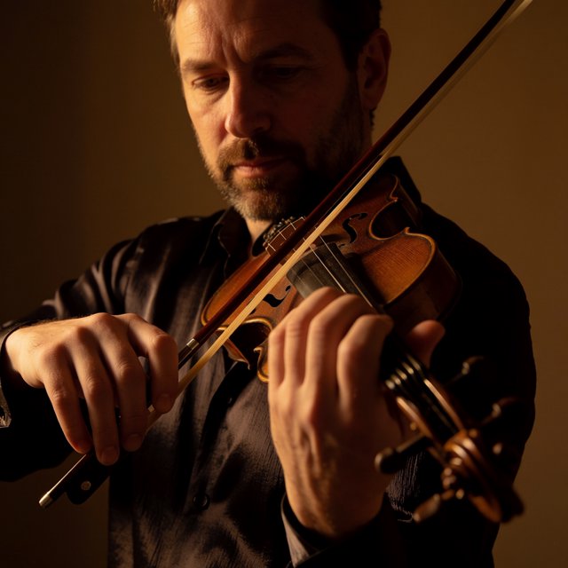 With a violin tucked close, an adult man in his early 50s draws a bow, his hands prominent, wearing a charcoal silk shirt; his intent expression is framed against a warm umber backdrop in soft amber window light, in a close portrait photograph. — Krea 2 Medium