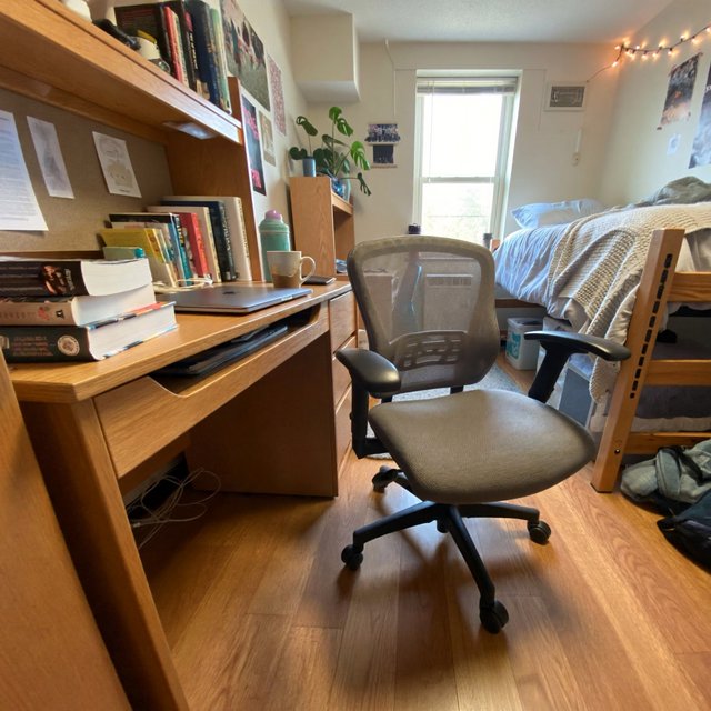 A dorm room belonging to an adult woman, photographed with a cell phone. The camera is sitting on a desk facing the empty chair and the rest of the room. — ImagineArt 2