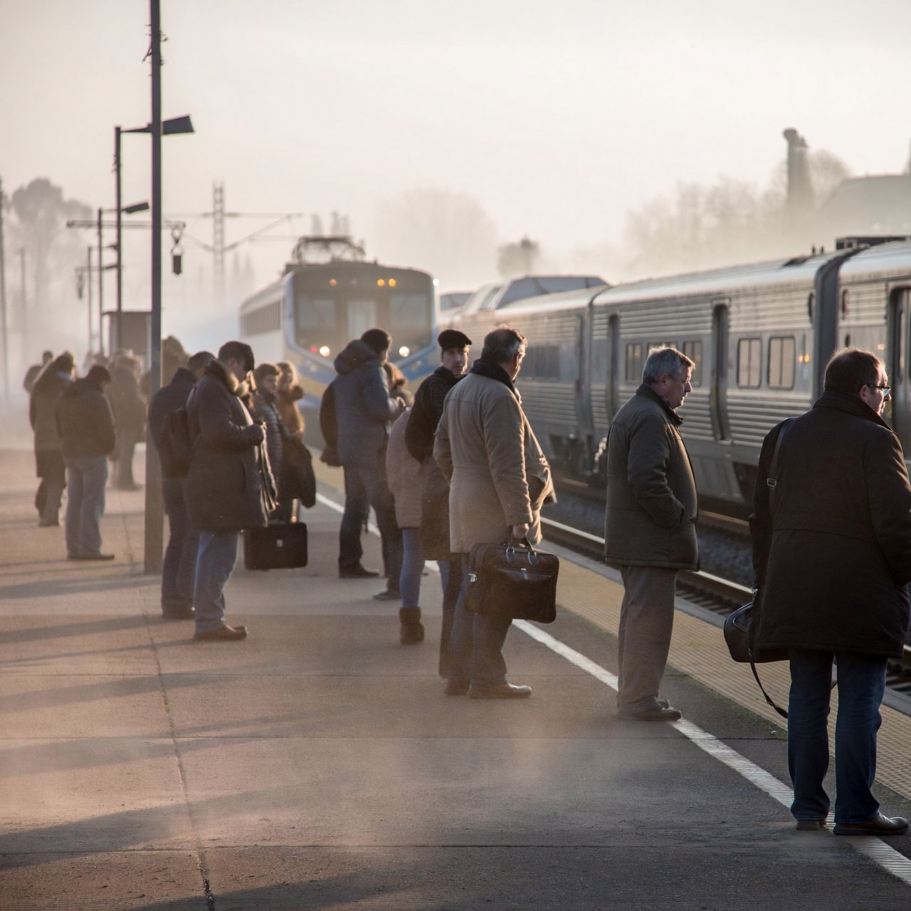 Across a broad station platform, adult commuters wait in loose clusters as a train approaches through pale dawn haze. A real-world press photograph uses a 135mm lens and deep focus; fine brake dust hangs in the sunlit air. — ImagineArt 2