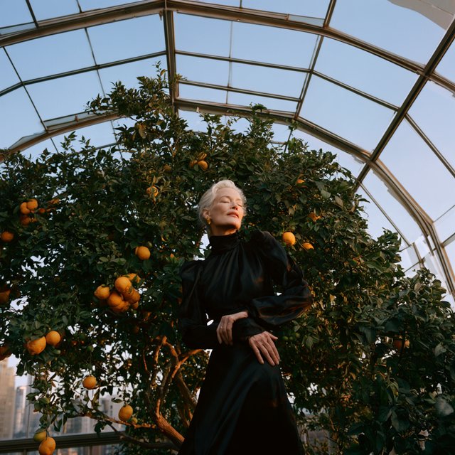 At a quiet rooftop conservatory, an adult woman in her early sixties wears a long-sleeved black evening gown, caught laughing softly beside a citrus tree as blue-hour light spills through the glass; poised styling brings fashion-magazine polish. — Model 13
