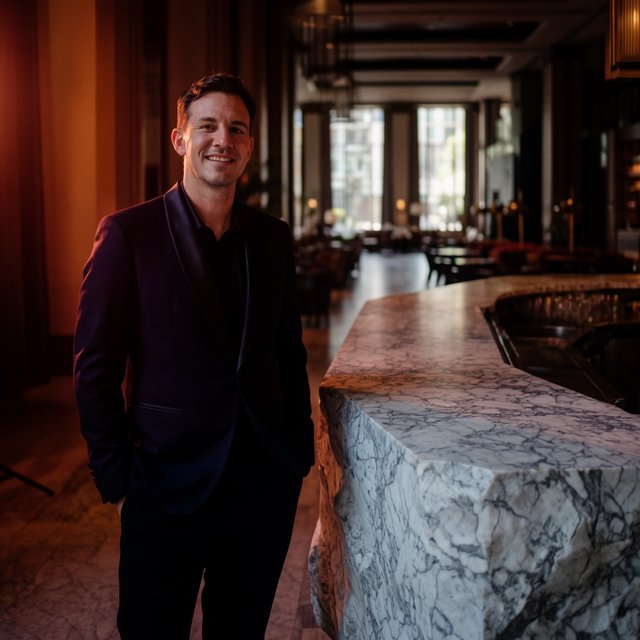 Editorial glamour photograph of an adult man in a midnight-plum dinner jacket and black trousers, standing naturally beside a sculptural marble bar in a quiet hotel lounge, flattering amber light, easy smile, polished editorial elegance. — Krea 2 Large