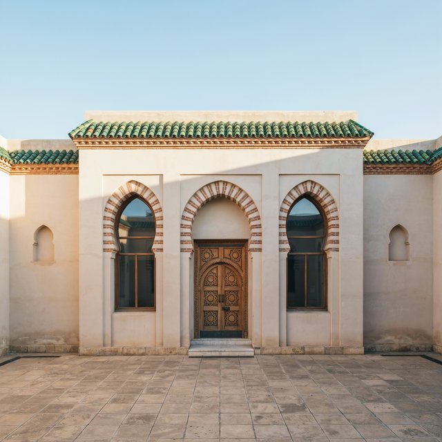 Exterior of a Moorish Revival municipal bathhouse, pale buff stucco, striped brick arches, green glazed tile roof and carved cedar doors; clear morning light, viewed across a paved courtyard, with coherent structure and crisp lines. — Ideogram v4.5