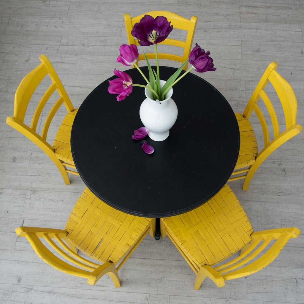 Exactly four yellow wooden chairs surround a round black table: two chairs at the front, one on the left, and one on the right. A white vase with three purple tulips sits at the table center, nothing else. — Grok Imagine Image Quality