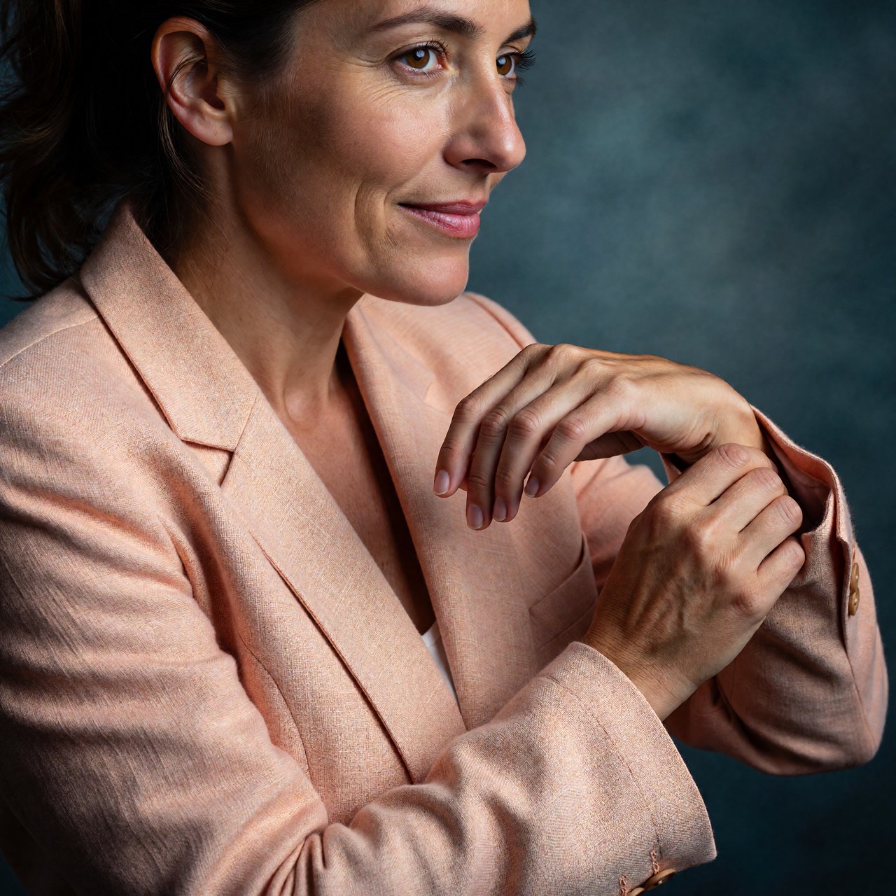 Portrait photograph of an adult woman in her early 30s wearing a pale apricot linen blazer, her hands gently smoothing its cuff. She looks quietly delighted against a deep teal paper backdrop, illuminated by soft, angled studio light. — ImagineArt 1.5 Pro