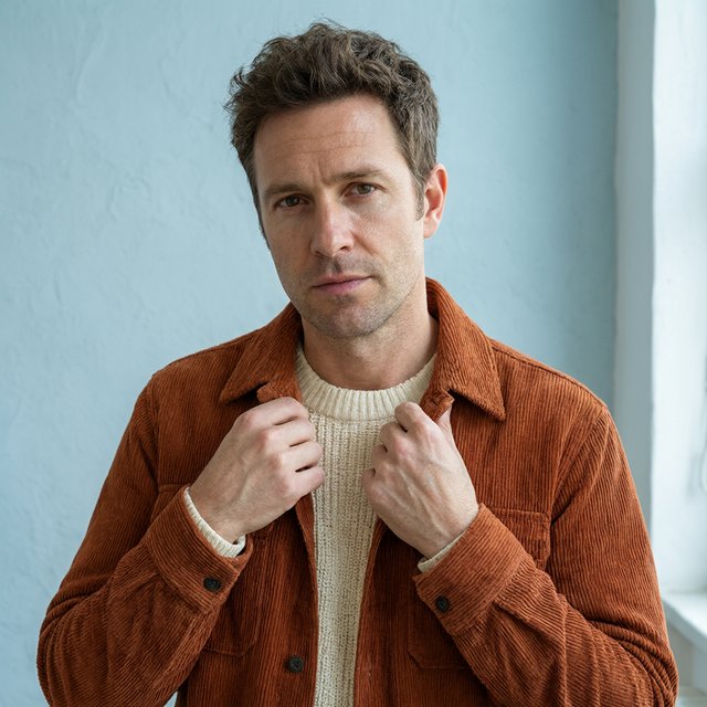 Portrait photograph of an adult man in his late 30s wearing a rust-colored corduroy overshirt over a cream knit, fingertips resting on its broad collar. His quietly curious gaze meets the camera against a pale blue plaster backdrop, softly lit by cool side-window light. — Qwen Image 3.0 Pro