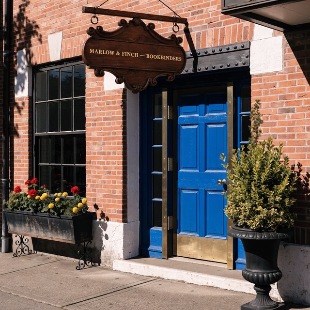 A hanging wooden shop sign that reads "MARLOW & FINCH — BOOKBINDERS" in gold serif letters, carved, above a blue door. — Recraft v4 Pro