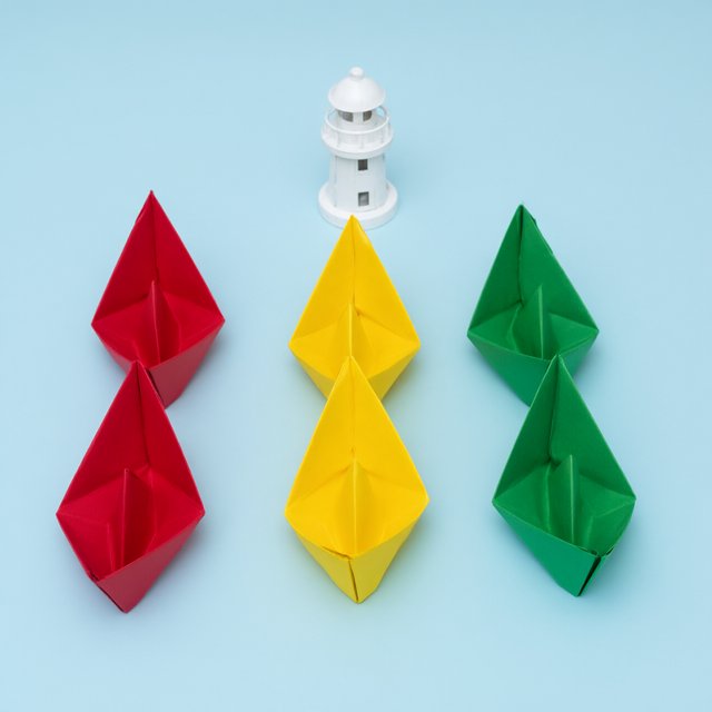 Exactly six folded paper boats sit on a pale blue tabletop: two red boats on the left, two yellow boats in the center, and two green boats on the right. A small white lighthouse figurine stands behind the center pair, nothing else. — Qwen Image 3.0