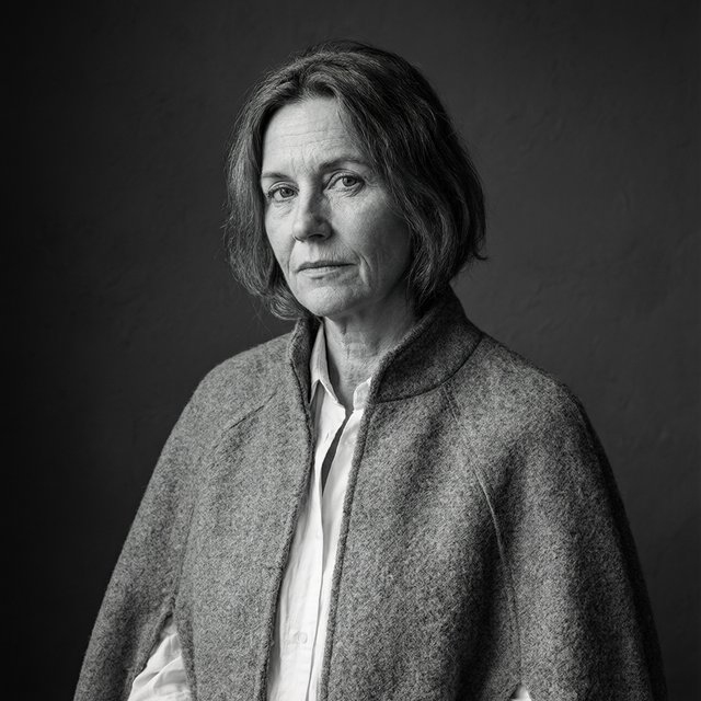 Under a narrow window beam, an adult woman in her late 50s wears a sculpted wool cape over a cotton blouse; her wry expression rests against a charcoal plaster backdrop in a black-and-white portrait, shaped by soft angled light. — Qwen Image 3.0 Pro