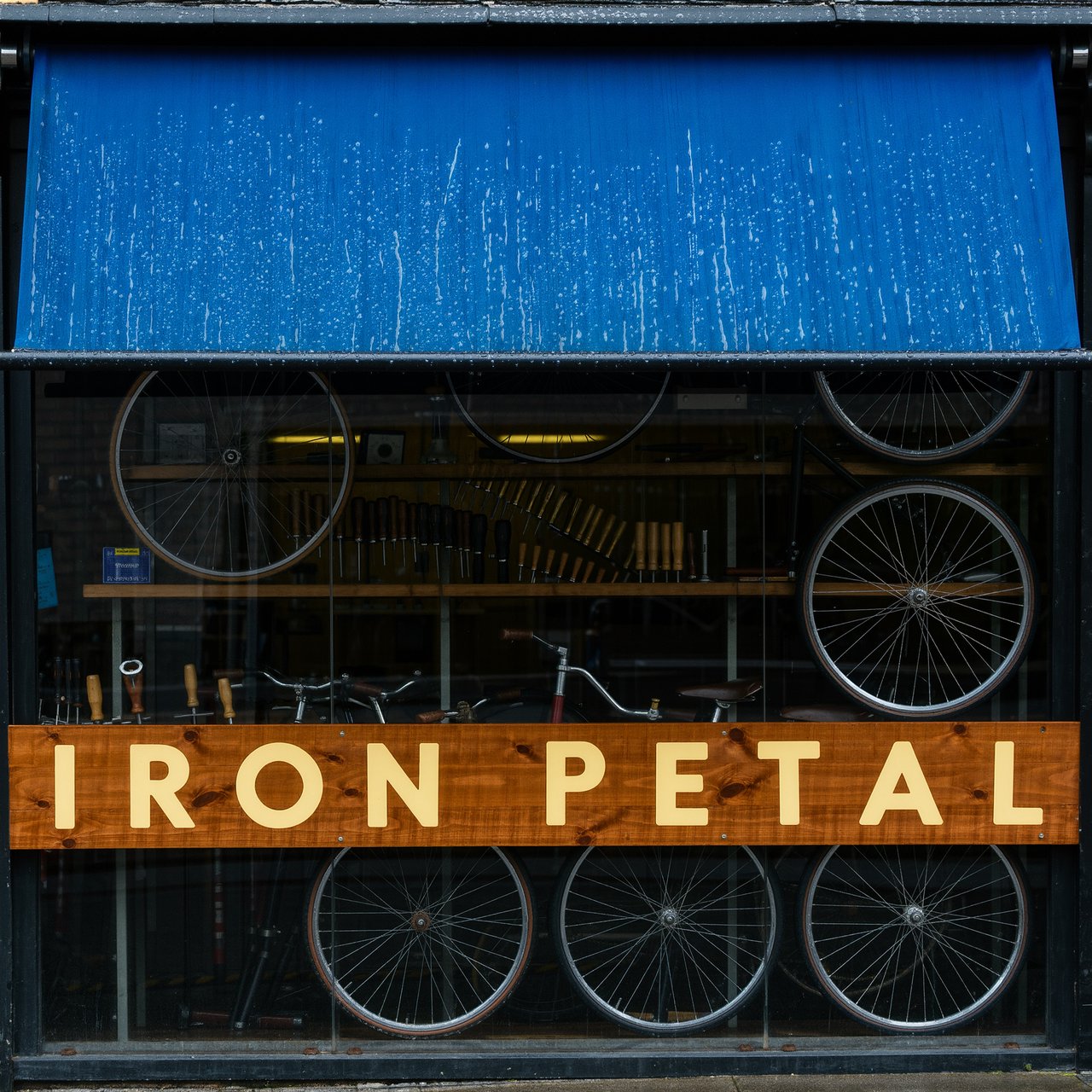 Rain beads on a bicycle repair shop’s blue awning, with wheels and brass tools visible behind the glass; “IRON PETAL” is lettered in warm yellow enamel capitals across the wooden fascia. — Kling v3 Omni