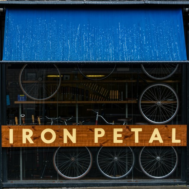 Rain beads on a bicycle repair shop’s blue awning, with wheels and brass tools visible behind the glass; “IRON PETAL” is lettered in warm yellow enamel capitals across the wooden fascia. — Kling v3 Omni
