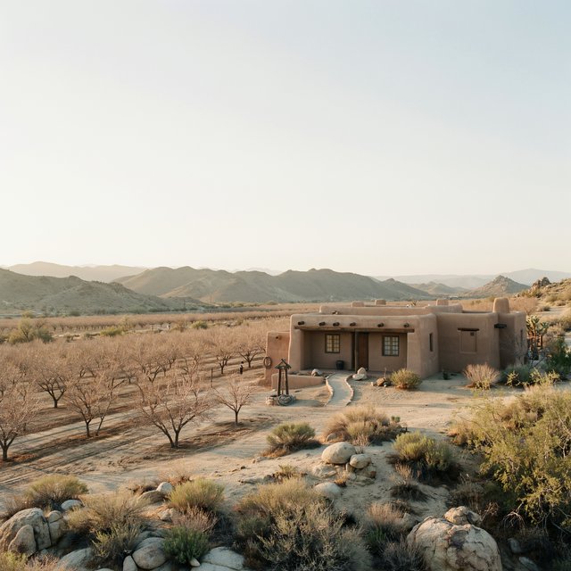 Beyond dry orchard rows, a Pueblo Revival adobe house settles into the desert foothills, with timber lintels and a muted clay roof; warm late-afternoon light, viewed from a rocky rise, reveals clean geometry and credible construction. — Ideogram v4
