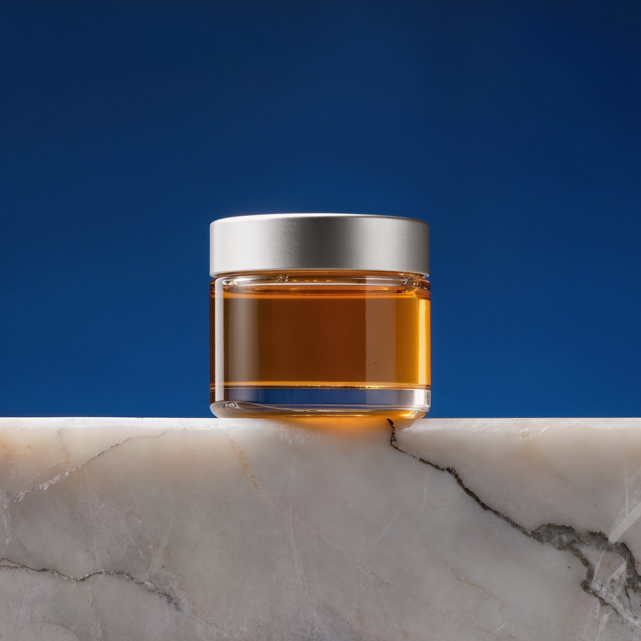 A squat unbranded honey jar with clear glass walls and a plain brushed-aluminum lid rests on honed pale limestone; broad diffused key light and warm rim light shape the commercial product image against a seamless cobalt-blue background, with no lettering. — Kling v3