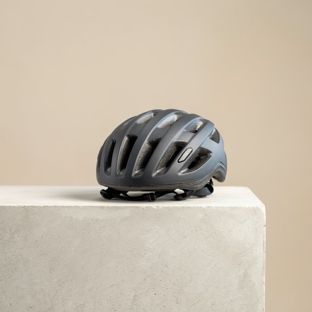 Commercial product photograph of an unbranded folding bicycle helmet made from matte charcoal polymer with soft gray padding, resting on a honed concrete plinth against a muted sand background, illuminated by a broad overhead softbox and gentle rim light. — Ideogram v4