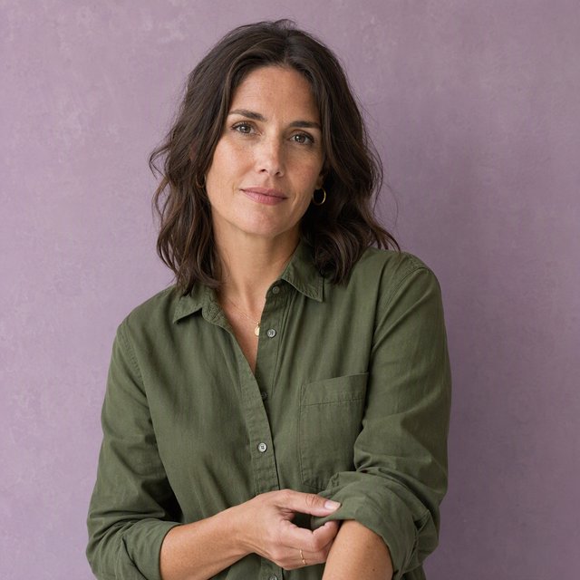 Portrait photograph of an adult woman in her late 30s wearing a moss-green cotton poplin shirt, fingers lightly turning its rolled cuff. Her calm, observant expression faces the camera against a chalky lavender backdrop, lit by soft diffused skylight. — GPT Image 2