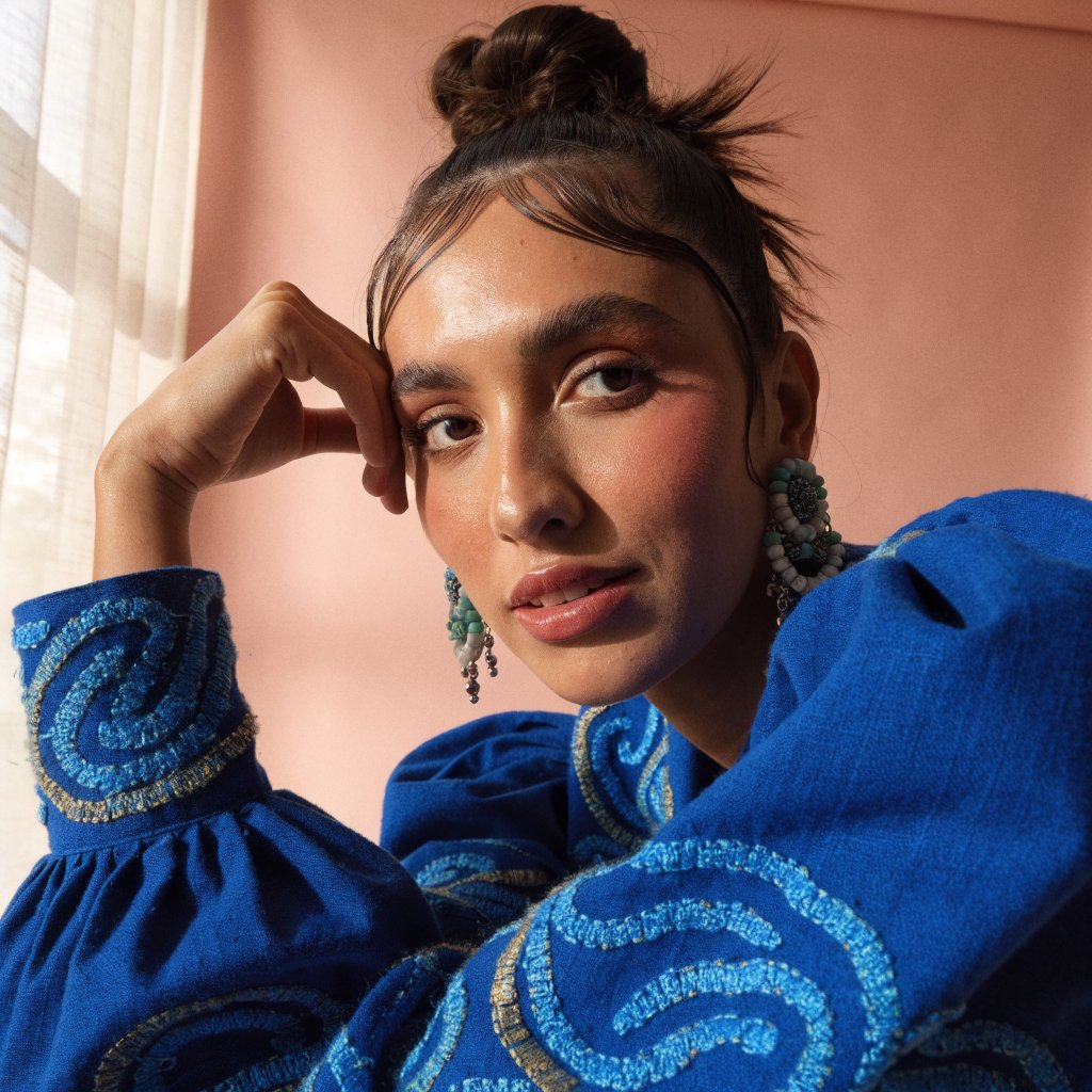 Fashion portrait of an adult Latina woman in her late 20s wearing a handwoven cobalt-blue cotton jacket, one hand touching its embroidered cuff, curious half-smile, against a pale peach studio backdrop, soft diffused window light shaping her face. — Recraft v4