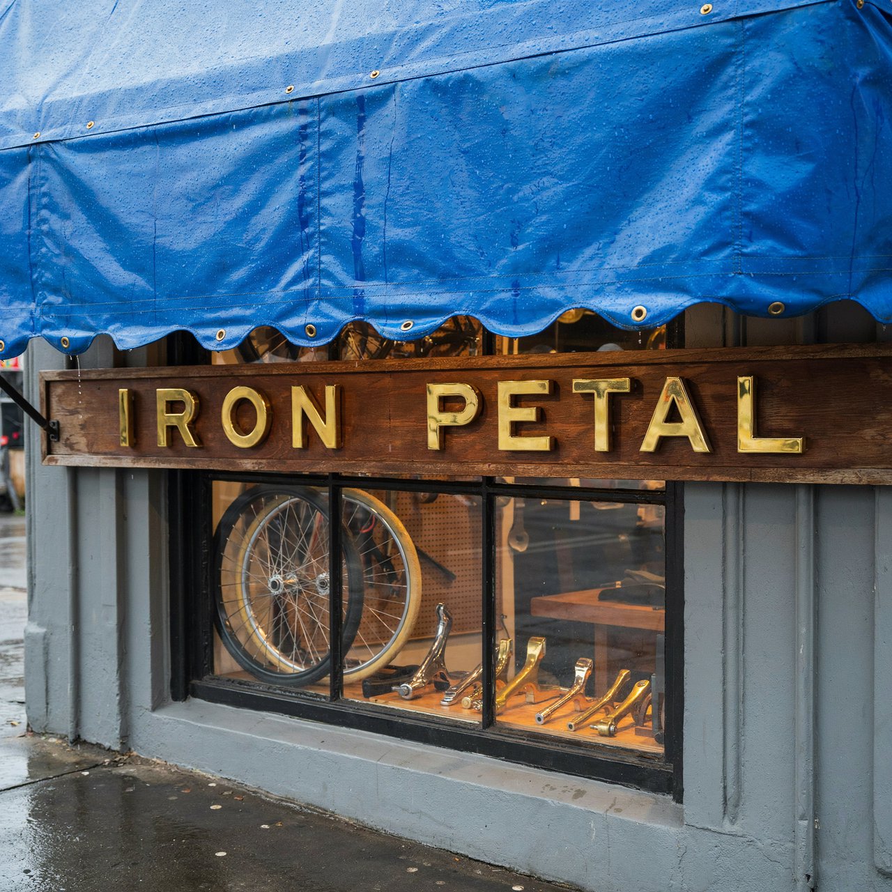 Rain beads on a bicycle repair shop’s blue awning, with wheels and brass tools visible behind the glass; “IRON PETAL” is lettered in warm yellow enamel capitals across the wooden fascia. — Ideogram v4.5