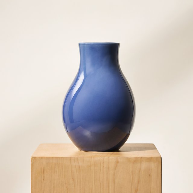 Commercial product photograph of an unbranded sculptural ceramic vase, formed from glossy cobalt porcelain, standing on a pale oak pedestal against a warm ivory background, illuminated by soft directional daylight and subtle fill. — Ideogram v4