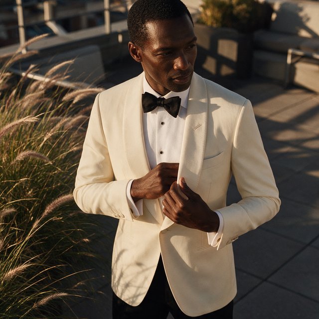 Under the copper glow of a rooftop terrace, an adult Black man in a tailored cream dinner suit is caught smoothing his lapel as evening breeze stirs nearby grasses; calm poise, sculpted shadows and refined fashion-magazine polish. — Seedream 5.0 Pro